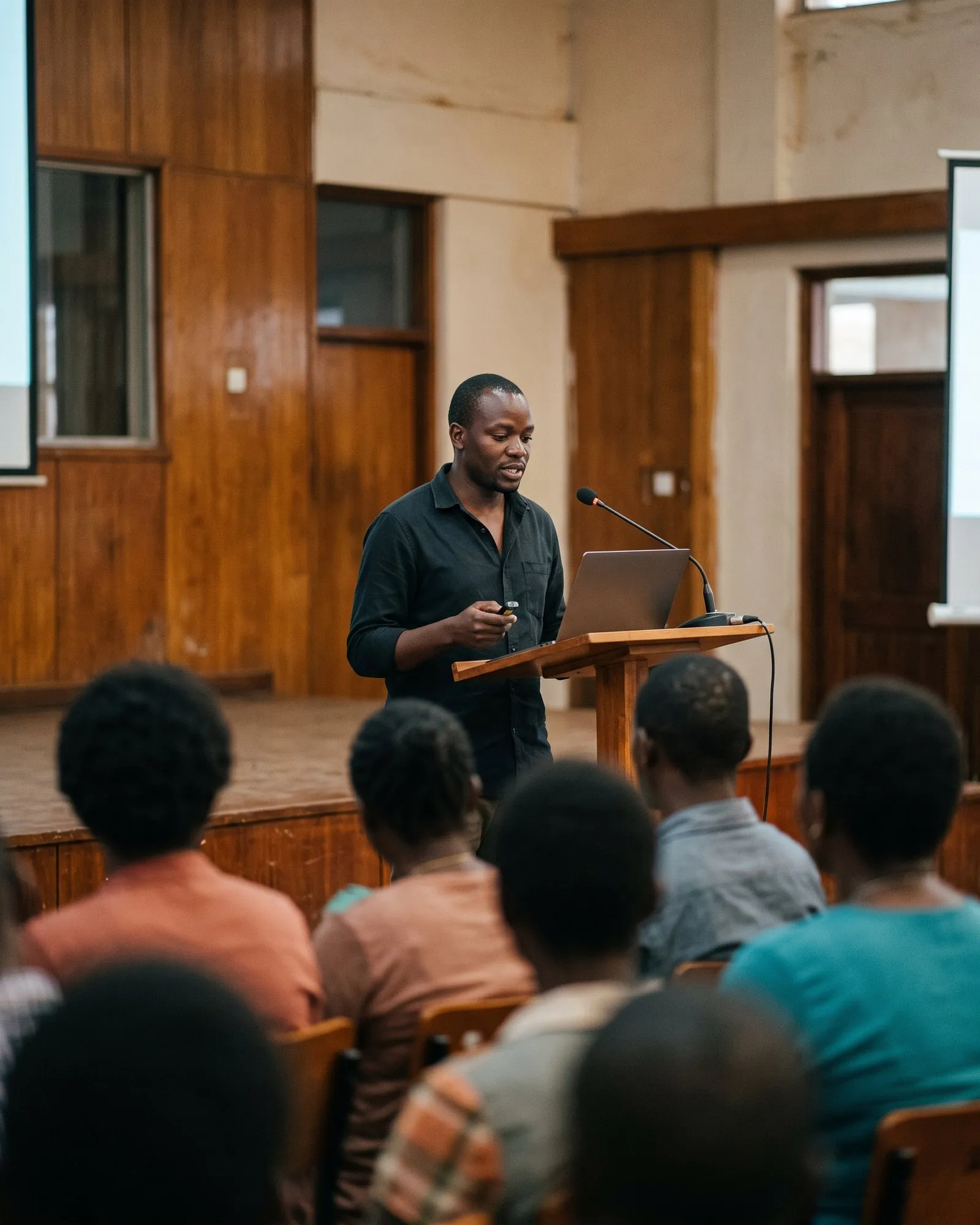 A presenter at a lectern glances at his laptop while speaking to a hall